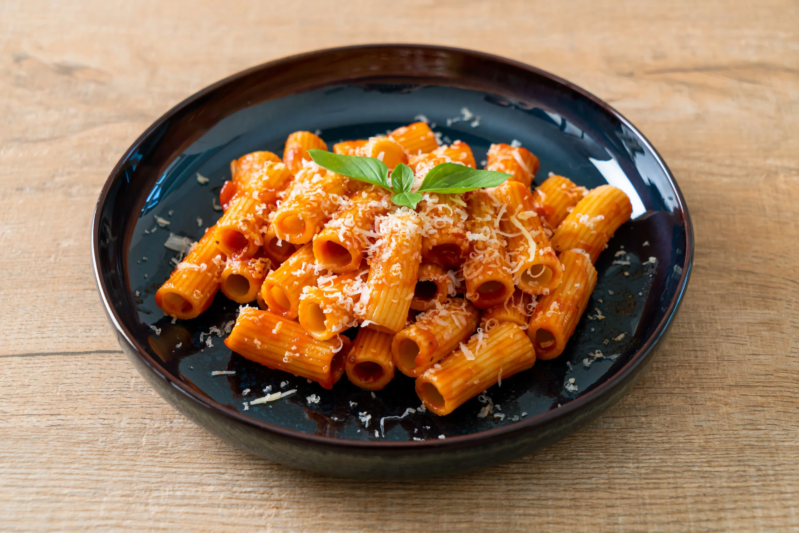 Ingredients for Carbone Spicy Rigatoni Recipe displayed on a rustic wooden surface, including rigatoni pasta, San Marzano tomatoes, Calabrian chili peppers, vodka, heavy cream, Parmesan cheese, garlic, shallots, fresh basil, olive oil, salt, and pepper.