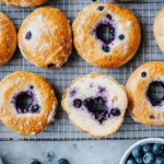 Freshly baked blueberry bagels with a golden crust, subtle blueberry specks, and a soft chewy texture, arranged on a white plate with cream cheese and fresh blueberrie